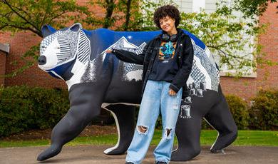 Anaisha stands next to a blue bear statue and points at it smiling 
