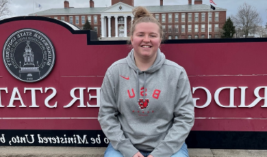 Addy Hodson wears a BSU sweatshirt sitting in front of red Bridgewater State University sign 